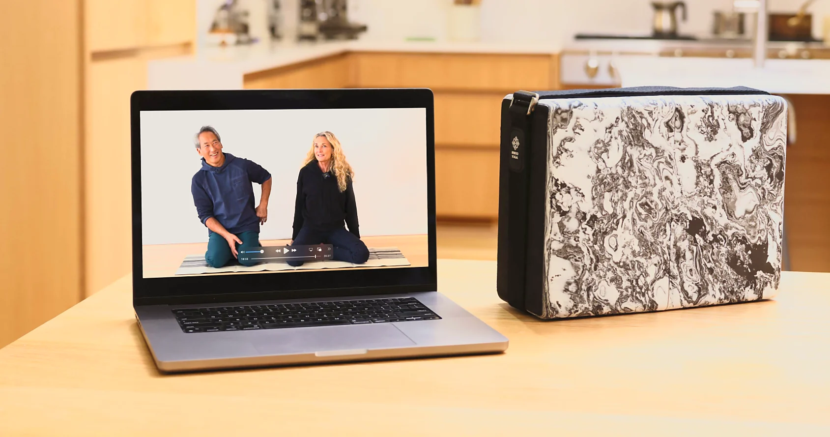 A laptop showing Rodney Yee and Colleen Saidman Yee teaching, resting on a kitchen counter beside the closed Bento Yoga ONE kit