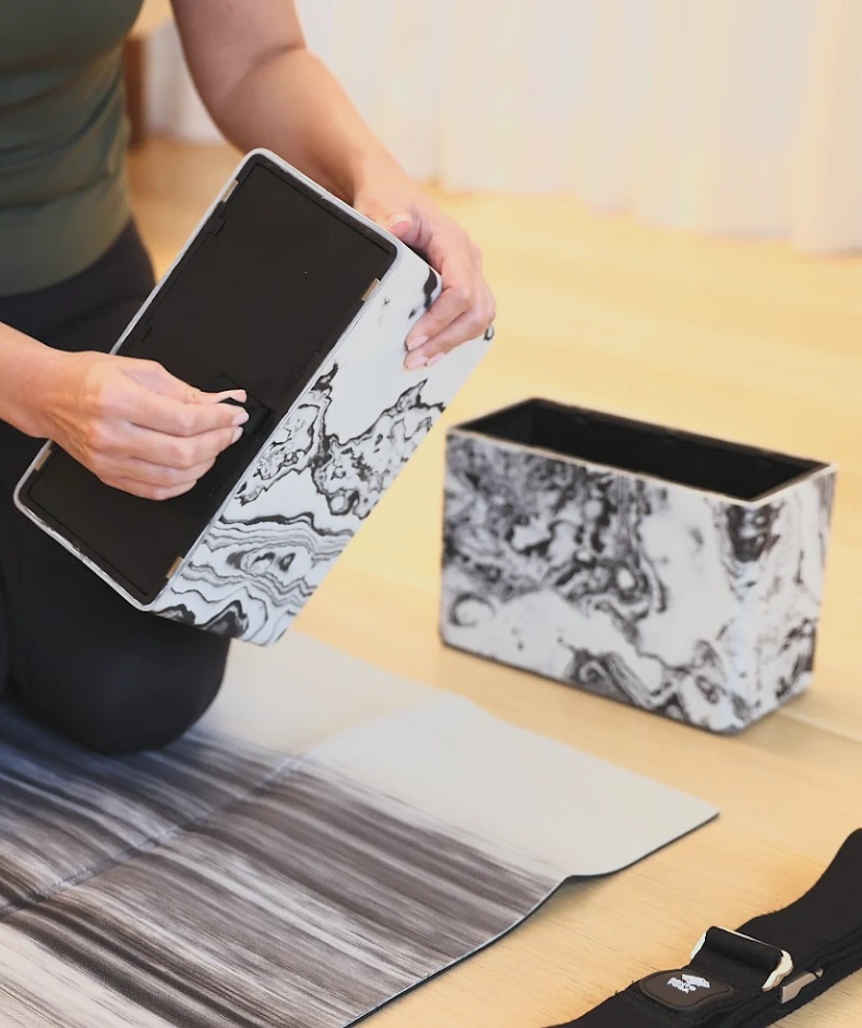 A practitioner closing the snap on a hollow Bento Yoga block, the folded mat and strap beside them